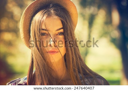 Beautiful young brunette woman with long hair flying in the wind and brown hat in park in summer. Head shot of gorgeous teenage girl with blue eyes. Retouched, filter, shallow depth of field.