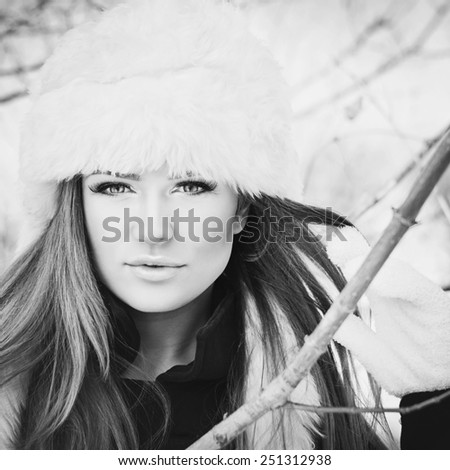 Gorgeous young Caucasian blonde woman in white fur hat and gloves posing outdoors in winter. Black and white, retouched, square image format, matte filter.