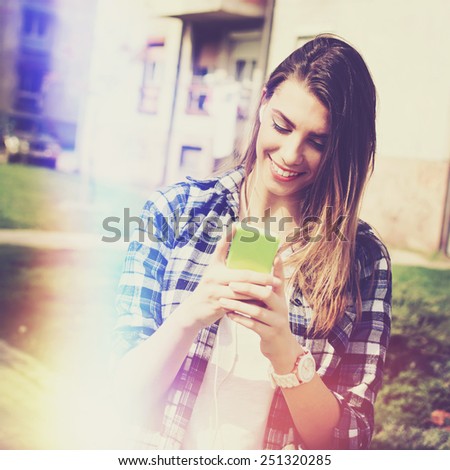 Teenage girl with smartphone and earphones smiling texting. Young Caucasian woman outdoors in summer with cellphone. Retouched, filter, light leak effect, vibrant colors, square format.