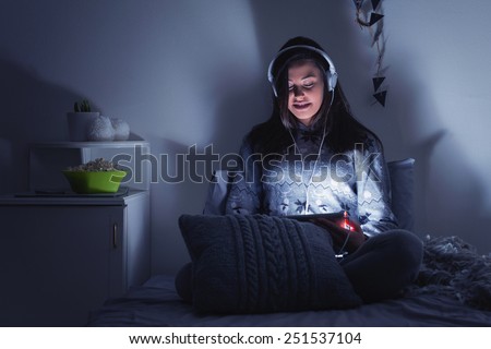 Teenage girl watching movie on tablet in bed at night. Happy brunette young woman with tablet and headphones using tablet at home in dark room. Horizontal, dark blue tones to emphasize night look.