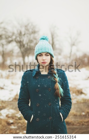 Beautiful young woman in green coat and mint beanie hat and dyed hair outdoors in winter. Hipster teenage girl in snow standing looking at camera. Retouched, filter, vertical, matte finish edit.
