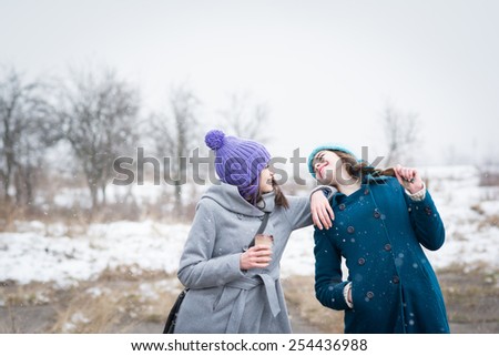 Two happy teenage girls with colorful knit beanie hats and colorful hair enjoying winter and snow outdoors. Young women in park with takeaway coffee laughing having fun on cold day. Mildly retouched.