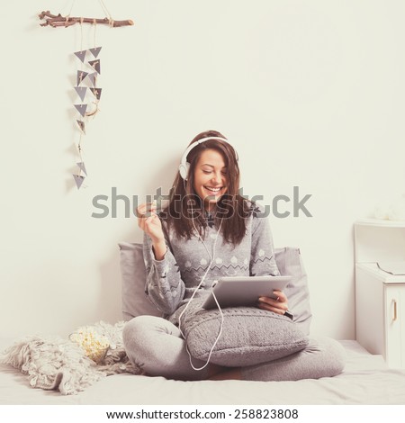 Beautiful happy young woman with headphones and tablet watching a movie on tablet in bed eating popcorn. Mild retouch, matte color filter, contemporary look.