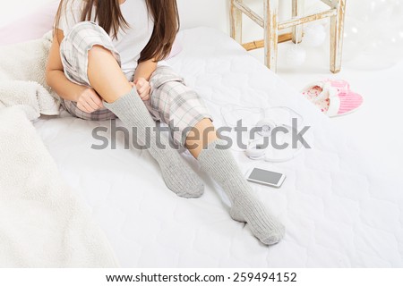 Closeup of young Caucasian brunette woman sitting in bed with socks and pajamas at home in the morning. Teenage girl with smartphone and headphones dressing. No retouch, horizontal.