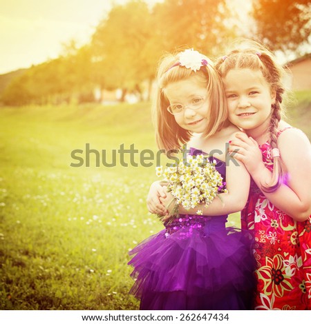 Two cute blonde little girls in park in spring. Two little blonde sisters in cute colorful dresses outdoors in summer smiling holding flowers bouquet. Retouched, square format.