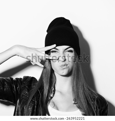 Fashionable hipster Caucasian brunette teenage girl with black beanie hat and black leather jacket posing showing two fingers and pouting. Medium retouch, black and white, square format.