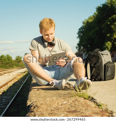 Young blond hipster man with digital tablet and headphones outdoors in summer sitting near railway on sunny summer day. Handsome teenage guy relaxing in park. Square format, mild retouch, no filter.