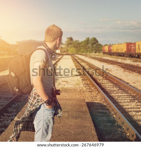Young Caucasian blond hipster man with backpack waiting for train at a station. Trendy casual guy standing looking away, unrecognizable person, square format image, filter applied.