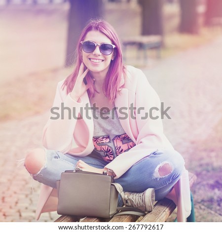 Trendy young woman with pink hair wearing fashionable denim jeans, pink coat and sunglasses smiling sitting on bench in park. Closeup, square format, retouched, color filter applied.