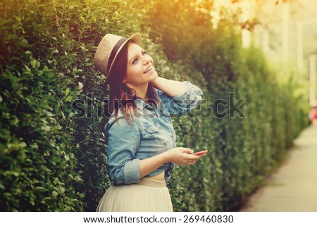 Boho styled young millennial redhead woman with smartphone in park on sunny summer day laughing. Modern girl with denim shirt, hat and beige skirt laughing enjoying. Horizontal, retouched. copy space.