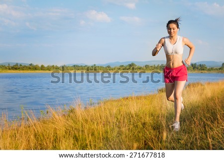 Fit young Caucasian brunette woman in short white tank top and red shorts running by the lake on sunny summer day. Horizontal, retouched, vibrant colors, natural light.