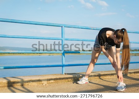 Young fitness Caucasian woman with long hair wearing black sportswear stretching after running outdoors on sunny summer day by the lake. Horizontal, mild retouch, natural light.