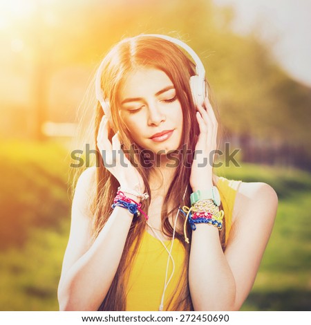 Young Caucasian brunette woman with headphones outdoors on sunny summer day. Millennial teenage girl listening to music wearing yellow shirt and vibrant jewelry. Square format, retouched, filter.