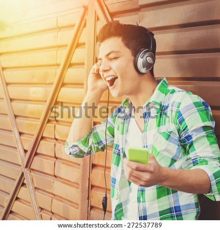 Hipster mixed race young man singing. Closeup of handsome guy with headphones and smartphone in green plaid shirt listening to music. Natural light, retouched, filter applied, square format.