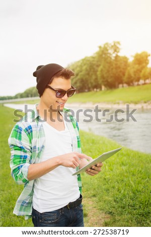 Hipster man with digital tablet by the river. Young mixed race hipster guy in casual clothes and sunglasses in park in summer. Closeup, vertical, retouched, vibrant colors.