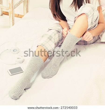Young Caucasian brunette woman in pajamas sitting in bed wearing soft gray socks, smart phone and headphones beside her. Square format, filter, unrecognizable person.