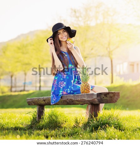 Cute happy bohemian young woman in blue floral dress and fedora hat sitting on a bench in park on sunny summer day. Portrait of boho teenage girl. Natural light, retouched, square format.