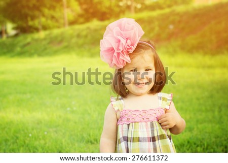 Cute little girl with big pink tulle flower headband smiling wearing green and pink dress standing outdoors in park on sunny summer day. Horizontal, copy space, retouched, vibrant colors.