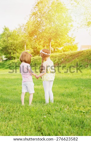 Two little sisters holding hands standing in park on sunny day. Rear view of two five year old little girls in pastel clothes outdoors in nature. Vertical, retouched, vibrant colors.