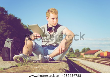 Handsome blond Caucasian young man with tablet, headphones and backpack sitting by the train rails looking away. Horizontal, retouched, filter applied.