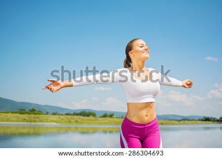 Smiling young blonde Caucasian woman in pink and white sportswear spreading hands feeling free standing by the lake. Fitness girl with hands wide stretched outdoors on sunny summer day.