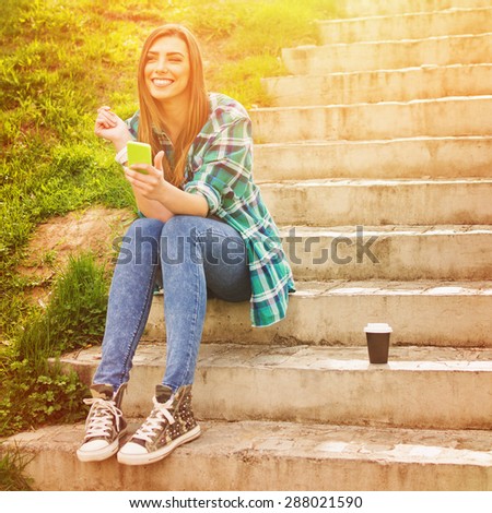 Happy beautiful teenage girl sitting on stairs in park on sunny summer day with smartphone and takeaway coffee. Square format, retouched, vibrant colors.