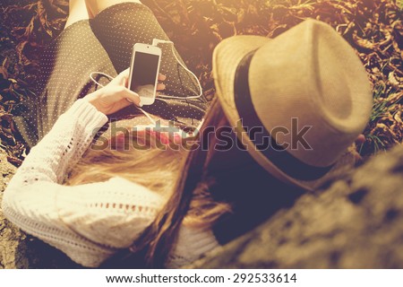 Closeup rear view of young woman in crochet blouse and skirt sitting next to a tree in autumn afternoon using smartphone texting. Horizontal, mild retouch, warm tones.