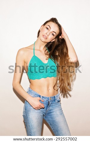 Gorgeous young woman with long blonde hair in turquoise crochet bikini top and blue denim jeans against white background. Vertical, retouched, studio shot, intentional shadow.