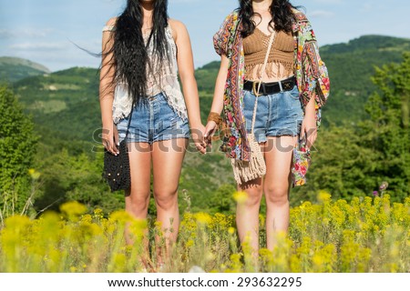 Two young hippie women holding hands standing outdoors in nature on sunny summer day. Two unrecognizable boho girls in park wearing bohemian outfit. Horizontal, no retouch, natural light.