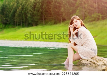 Beautiful young blonde woman at the beach sitting on the stone dipping legs in water looking at camera posing wearing sheer beige embroidered dress. Horizontal, natural light, retouched.