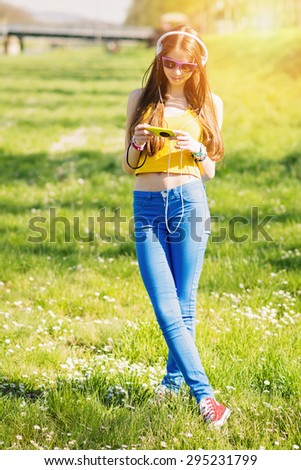 Modern teenage girl in yellow crop top, blue pants and red canvas shoes with smartphone and headphones smiling standing in park on sunny summer day. Vertical, retouched, vibrant colors.