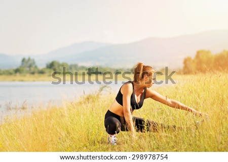 Fit young woman in black tank top and leggings stretching outdoors by the lake on sunny summer day. Retouched, vibrant colors, horizontal.