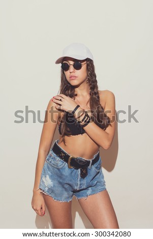 Trendy boho young woman in crochet bikini top, denim shorts, white cap and sunglasses. Modern brunette in bohemian summer outfit with braids. Matte filter, studio shot, intentional shadow on the wall.