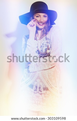 Young woman with blonde hair in black fedora hat and boho sheer lace crochet beige dress posing against white wall making mustache with her hair. Color filters applied, intentional light leak effects.