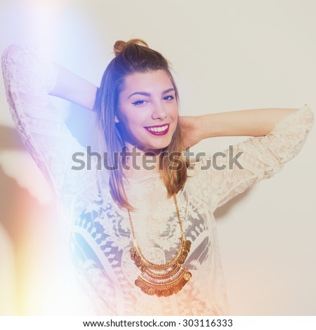 Young woman with blonde hair in boho sheer lace crochet beige dress and golden necklace posing against white wall smiling looking at camera Color filters applied, intentional light leak effects.