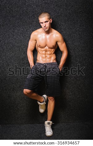 Fit muscular young shirtless man posing. Young Caucasian bodybuilder guy with short hair standing against black background wearing gray shorts. Medium retouch, no filter.