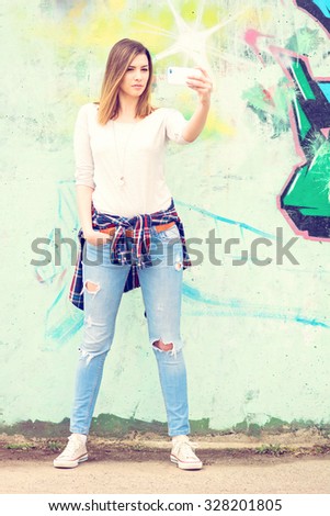 Fashionable young blonde woman in white blouse and denim jeans taking a selfie against colorful wall outdoors. Teenage girl photographing herself on smartphone. Vertical, pastel colors, mild retouch.