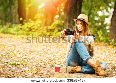 Modern young millennial woman taking a selfie with digital camera.  Teenage girl with hat, camera, takeaway coffee photographing herself sitting outdoors in autumn. Retouched, vibrant colors.