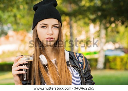 Beautiful young woman with beer or coffee in park. Teenage girl in black beanie hat, leather jacket and headphones outdoors in autumn. Horizontal, mild retouch, vibrant colors.