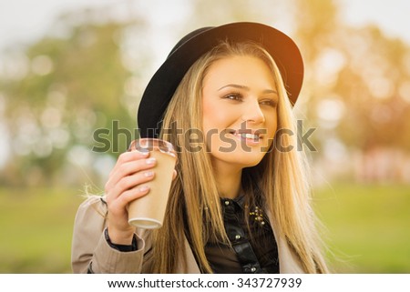 Happy young blonde Caucasian woman in park in autumn wearing trench coat and fedora hat holding a cup of takeaway coffee. Horizontal, retouched, vibrant colors.