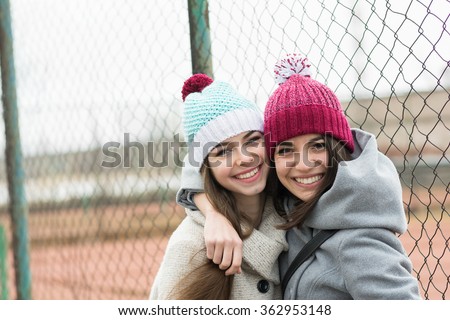 Two happy beautiful teenage girls in knitted beanie hats hugging. Two girlfriends without makeup outdoors in winter smiling and posing. Horizontal, no retouch, no filter, natural light.