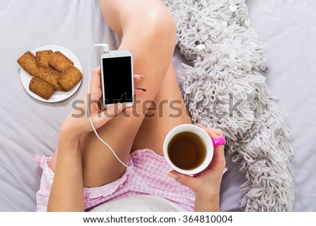 Closeup of smartphone and coffee held by young woman sitting in bed wearing short pink baby doll pajamas. Plate with cookies and gray knitted sweater next to her, horizontal, medium retouch.