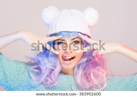Modern young woman with pastel blue and pink hair, wearing pastel green sweater and white beanie hat with pom poms posing, making funny facial expression. Retouched, studio portrait.