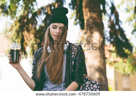Modern teenage hipster girl in autumn in park in beanie hat, with takeaway coffee and headphones Millennial young woman in fall relaxing, drinking cappuccino. Matte filter, natural lighting.