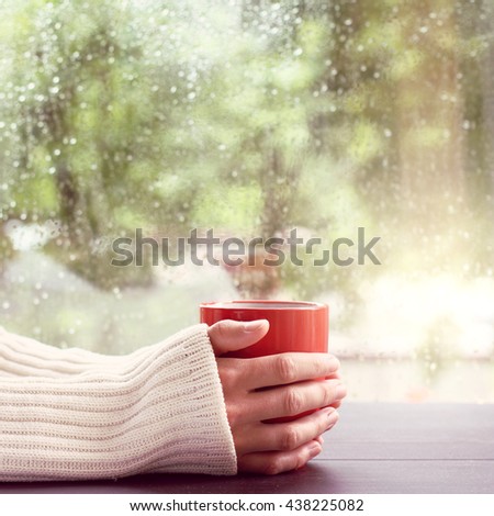 man in a light jumper, holds in hands red mug on a background of a wet window after the rain / hot drink and watch the rain