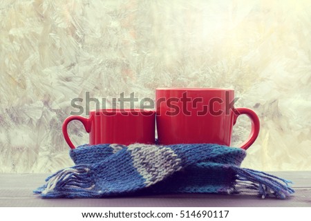 small and big red mug wrapped in a blue scarf and standing on the table against the background of a winter window / warming atmosphere for hanging out
