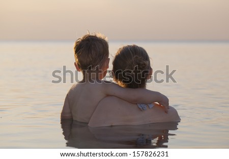 Mother and her little son standing chest high in a calm sea watching the sunset with their backs to the camera
