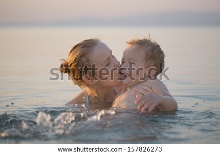 Mother playing in the sea with her little son splashing the surface and kissing him tenderly on the cheek as he laughs with delight