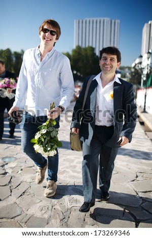 Groom and his friend - two smiling men walking down a street one carrying flowers and the other in a suit wearing a buttonhole flower