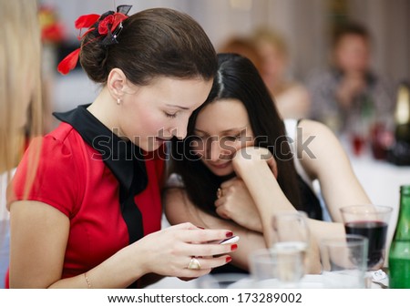 Two beautiful stylish young women at a formal event sitting together at a table reading an sms on a mobile phone